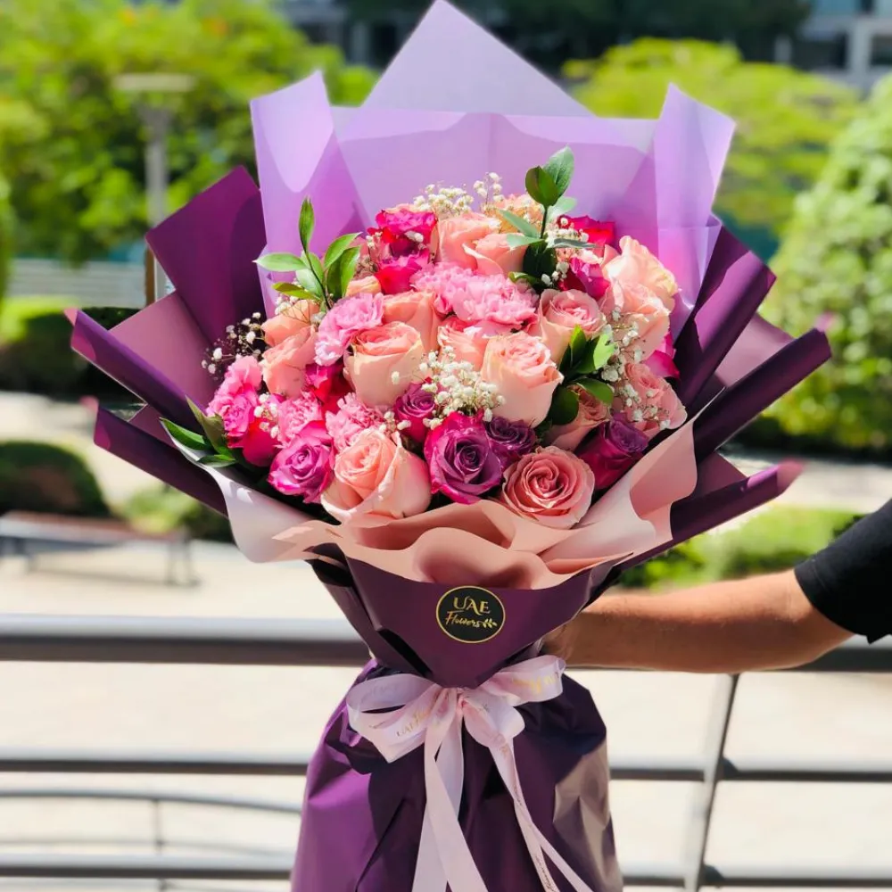 A bouquet of 9 light pink roses, 6 purple roses & 3 light pink carnations with fillers in a purple and grey wrapping paper tied with grey ribbon