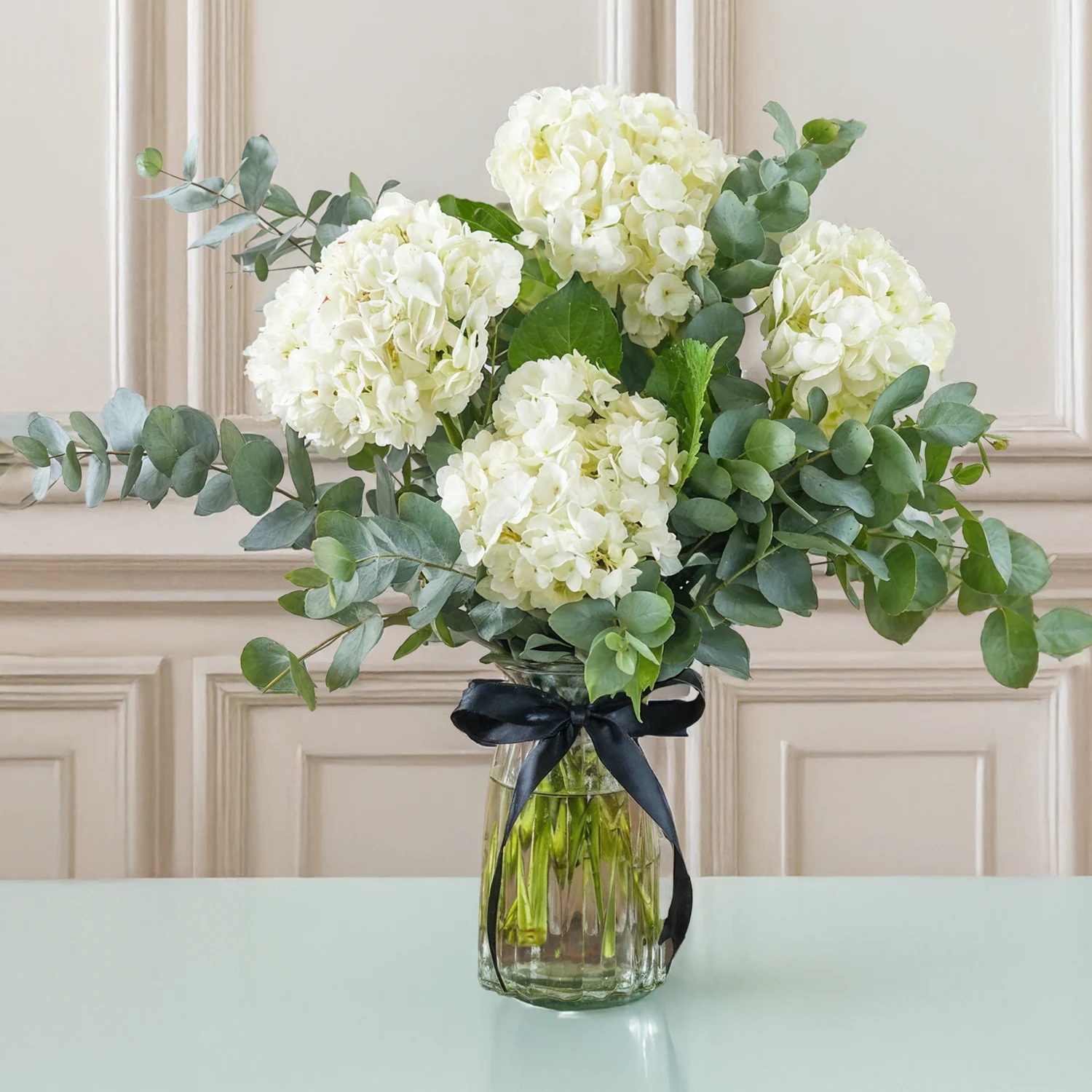 ouquet of 4 white hydrangeas with fresh eucalyptus leaves in a glass vase tied with a black ribbon.